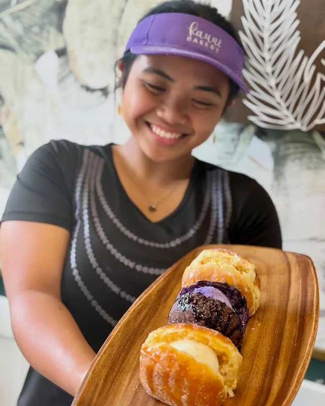 A Kauai Bakery team member in a purple cap holding a board of malasadas